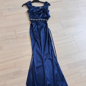 Marinblå festklänning - Elegant marinblå långklänning i glansigt satintyg med markerad midja och pärlbroderier vid midjebandet. Pärlorna är insydda i själva klänningen men har även band där bak man kan knyta. Klänningen har breda axelband, draperad överdel. Perfekt för bal eller festliga tillfällen. Använd 1 gång. Storlek xs.