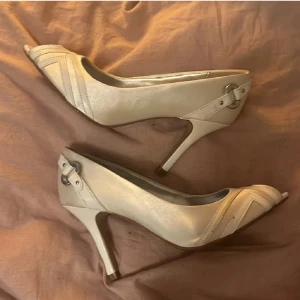 Vita klackar med öppen tå från Graceland - Snygga vita pumps från Graceland i satinliknande material med öppen tå och hög klack. Skorna har dekorativa sömmar och metallspänne vid hälen för en extra detalj. Perfekta för bal,fest och till sommarn. Några mindre defekter på ena skon (se bild). Fråga gärna❤️