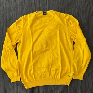 Gul stickad tröja från Hugo Boss - Snygg gul stickad tröja från Hugo Boss i italienskt garn. Tröjan har rund halsringning, långa ärmar och en liten metallbricka med logga vid nederkanten. Perfekt för dig som vill sticka ut med färg och stil.