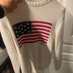 Vit stickad tröja Polo Ralph Lauren - Vit stickad tröja från Polo Ralph Lauren med amerikansk flagga i rött, blått och vitt på bröstet samt RL-broderi. Klassisk rund halsringning och ribbade muddar. Perfekt för dig som gillar clasic stil.