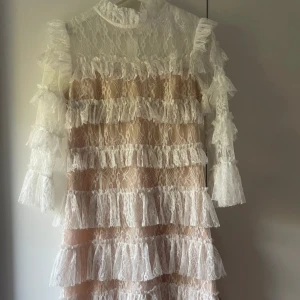 Vit spetsklänning från By Malina - Superfin vit spetsklänning från By Malina med volanger och hög hals Perfekt för dig som vill sticka ut med en unik och elegant stil.