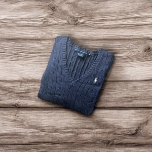 Mörkblå kabelstickad tröja Ralph Lauren - Säljer en mörkblå kabelstickad tröja från Ralph Lauren med klassisk v-ringning och broderad logga på bröstet. Tröjan har lång ärm och ribbade muddar, perfekt för lager-på-lager. Materialet är mjuk bomull som känns skönt mot huden.