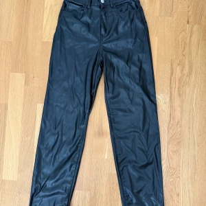 Svarta skinnbyxor från H&M Divided - Snygga svarta byxor i fejkskinn från H&M Divided. Klassisk femficksmodell med raka ben och normal passform. Perfekta för dig som vill ha en edgy look med lite glansig finish. Stängs med knapp och dragkedja framtill.