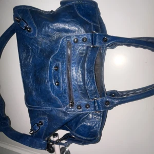 Blå skinnväska med dragkedja - Cool blå handväska i skinn med snygga detaljer och silverfärgade nitar. Väskan har dubbla handtag, dragkedja framtill och ett rymligt innerfack. Perfekt för dig som vill ha en statementväska med edgy vibe och plats för allt du behöver.