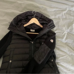 Svart Moncler pufferjacka med huva - Säljer en svart pufferjacka från Moncler med stickade ärmar och huva. Jackan har dragkedja framtill, två fickor med tryckknappar och en Moncler-logga på ärmen. Perfekt för kyliga dagar och riktigt snygg till streetwear-stilen.