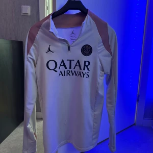 PSG långärmad träningströja Jordan - Snygg långärmad fotbollströja från PSG och Jordan med halv dragkedja. Tröjan är vit med svarta detaljer, PSG-emblem på bröstet och Jordan-logga. Tillverkad i Dri-FIT-material som andas, perfekt för träning eller match. Storlek är S men passar som M