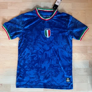 Italien blå fotbollströja Calcio Italia - Blå fotbollströja med konstnärligt mönster inspirerat av italiensk renässans. Tröjan har korta ärmar och ribbade muddar i Italiens flaggfärger. På bröstet finns Italiens sköld med tre stjärnor. Materialet är lätt och andas, perfekt för match eller träning.