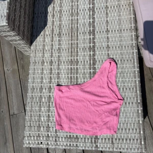 Rosa one shoulder topp från ASOS - Säljer en ribbad rosa one shoulder topp från ASOS DESIGN. Toppen är croppad och har en snygg asymmetrisk skärning med en axel. Perfekt för dig som vill sticka ut med en färgglad och trendig look. i storlek L. Passar m/l