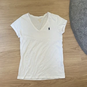 Vit v-ringad t-shirt från Polo Ralph Lauren - Vit t-shirt från Polo Ralph Lauren med v-ringning och klassisk marinblå logga broderad på bröstet. T-shirten är kortärmad och har en slim passform, perfekt för en clean och stilren look.