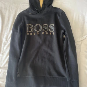 Svart hoodie från Hugo Boss med logga - Svart hoodie från Hugo Boss med stor guldfärgad BOSS-logga på bröstet och guldfärgad insida på huvan. Snygga detaljer med BOSS-tryck längs sidorna. Klassisk passform, långärmad och med dragsko i huvan. Perfekt för en stilren och trendig look.