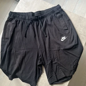 Svarta Nike shorts med snörning - Svarta shorts från Nike med vit logga på vänster ben. Mjukt bomullsmaterial, elastisk midja med snörning och praktiska fickor fram samt en bakficka. Perfekta för chill dagar eller träning. Klassisk loose fit och enkel stil som funkar till allt. Skick 8/10.