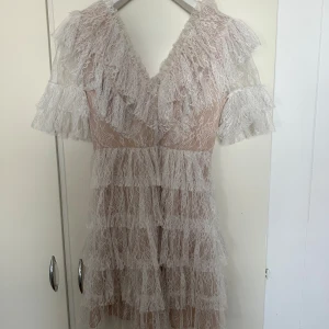 Ljus spetsklänning från Malina - Superfin kortklänning från Malina i ljusbeige med flera lager volangig spets. Djup v-ringning, korta ärmar och romantisk känsla. Perfekt för dig som gillar detaljer och en feminin look.