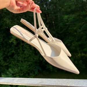 Vita ballerinaskor med spetsig tå  - Snygga ballerinaskor/sandaler/pumps från Nelly med spetsig tå och tunna remmar runt hälen. Skorna har platt sula och är tillverkade i ett syntetmaterial som ger en stilren och minimalistisk look. Perfekta för dig som gillar en clean och trendig stil. Storlek 39! 