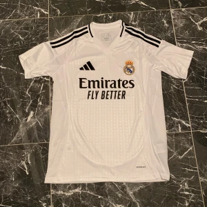 Real Madrid vit matchtröja Adidas - Snygg vit Real Madrid fotbollströja från Adidas med svarta detaljer och klubbmärke på bröstet. Tröjan har korta ärmar,  och är tillverkad i lätt, ventilerande funktionsmaterial. Klassiska svarta ränder på axlarna och tryck med Emirates Fly Better på framsidan.
