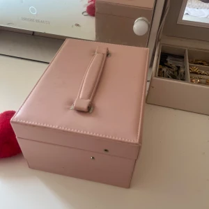 Ljusrosa smyckesbox med spegel - Söt ljusrosa smyckesbox i syntetmaterial med handtag på locket och inbyggd spegel. Boxen har flera fack för ringar, örhängen och andra smycken samt en utdragbar låda. Perfekt för att hålla ordning på dina favoritsmycken.
