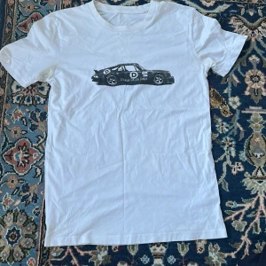 Vit t-shirt med biltryck - D’once De La Sense tröja med tryck som föreställer en Porsche på bröstet. Tröjan är använd men är ändå i fint skick. Denna tröja är perfekt för sommaren. Skriv vid frågor!