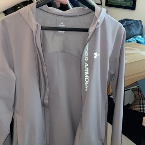 Grå vindjacka från Under Armour - Snygg grå vindjacka från Under Armour med huva och dragkedja. Jackan har logga och text i vitt och grönt framtill, samt ventilerande mesh på insidan. Perfekt för träning eller när du vill ha en sportig look. Storlek S, otroligt välskött och bra skick 