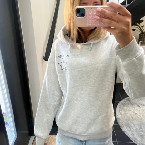 Grå hoodie med stjärna och text - Ljusgrå hoodie med texten Ma Chérie med svart broderad stjärna och text både fram och bak. Tröjan har huva med snörning och ribbade muddar vid ärmslut och nederkant. Perfekt för chill dagar och enkel att matcha med jeans eller kjol på sommaren