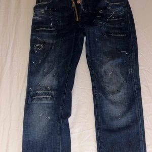 Dsquared2 blå jeans - Säljer ett par blå jeans från Dsquared2 med coola slitningar, färgstänk och trasiga detaljer. Jeansen har raka ben, klassisk femficksmodell och dragkedja framtill. Perfekta för dig som gillar en edgy streetstil. OBS! Pris kan diskuteras