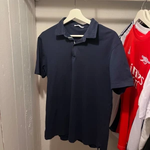 Mörkblå pikétröja från Zara - Säljer en stilren mörkblå pikétröja från Zara i storlek S. Tröjan har klassisk krage, korta ärmar och är tillverkad i mjuk bomull. Perfekt för en clean och avslappnad look.