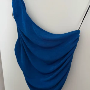 Blå draperad topp från Pieces - Snygg blå topp från Pieces i storlek L. Toppen har en draperad design med tunna axelband och en cool, asymmetrisk form. Perfekt för dig som vill sticka ut med en färgstark look. Materialet känns mjukt och följsamt mot huden.