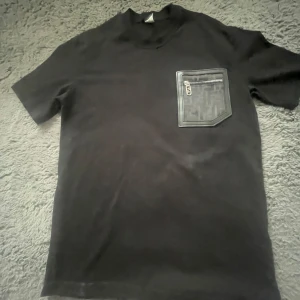 Svart Fendi t-shirt med ficka och dragkedja - Svart t-shirt från Fendi med korta ärmar och en unik bröstficka med dragkedja och Fendi-logomönster. Fickan har en snygg läderliknande kant och detaljerad zip. Perfekt för dig som gillar exklusiva streetwear-plagg med lyxig känsla. Den ser fläckig ut men den är inte de!