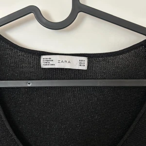 Svart glittrig klänning från Zara - Supersnygg svart klänning från Zara med glittrig finish. Klänningen har lång ärm, rundad urringning och ett lätt utställt kjolparti. Materialet är en mix av polyester och metalltråd som ger en skimrande look. Perfekt för dig som vill sticka ut lite extra.
