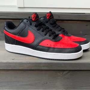 Nike Court Vision Low svart/röd - Snygga Nike Court Vision Low sneakers i svart och rött skinn med klassisk vit sula. Skorna har perforerad tå, svart snörning och röd swoosh på sidorna. Perfekt för dig som gillar retrovibbar och streetstyle.