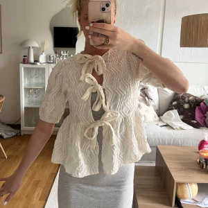 Krämvit blus med knyt fram - Supersöt krämvit blus med struktur och tre knyt framtill. Blusen har korta ärmar och volang nedtill, perfekt för en chill och trendig look. Materialet känns luftigt och lätt, och den har en unik textur som sticker ut.