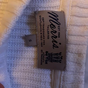 Vit stickad tröja från Morris - Vit stickad tröja från Morris med broderad Union Jack-flagga och texten 'Morris Great Britain' på bröstet. Tröjan har ribbade muddar, rund hals och lång ärm. Perfekt för dig som gillar klassisk brittisk stil med en modern twist.