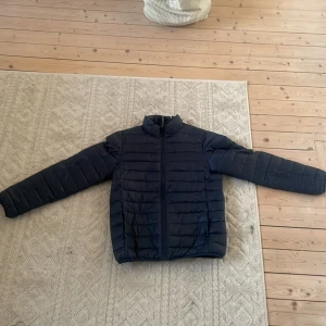 Svart pufferjacka med dragkedja - Säljer en svart pufferjacka med horisontella sömmar och hög krage. Jackan har en clean look med hel dragkedja framtill och långa ärmar. Perfekt för kyliga dagar när du vill hålla värmen utan att tumma på stilen.