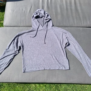 Grå ribbad tunn hoodie med huva - Säljer en grå ribbad hoodie med huva och snörning. Hoodien är väldigt tunn i materialet. Tröjan är croppad och har långa ärmar. Perfekt för dig som gillar en avslappnad och trendig stil. Mjuk och skön att bära, passar till många olika outfits.
