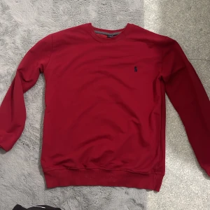 Röd tröja från Polo Ralph Lauren M - Röd långärmad tröja från Polo Ralph Lauren i storlek M. Klassisk rund halsringning och marinblå broderad logga på bröstet. Tröjan har ribbade muddar och är tillverkad i mjuk bomull, perfekt för en clean och stilren look.