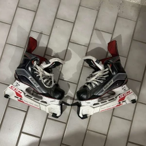 Svarta Bauer hockeyskridskor - Grymma svarta hockeyskridskor från Bauer med vita och röda detaljer. Klassisk snörning och robust design, perfekt för isen. Yttermaterial i syntet och insida i mjukt tyg för extra komfort.