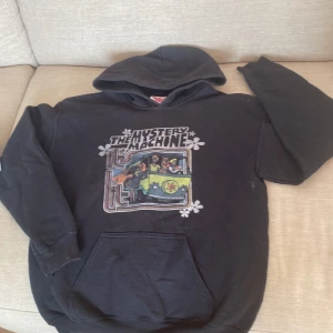 Svart Scooby-Doo hoodie - Svart hoodie med stor färgglad Mystery Machine-tryck från Scooby-Doo på bröstet. Tröjan har huva och känguruficka framtill. Perfekt för dig som gillar retro vibes och tecknade serier. Mjuk och skön, passar till chill dagar.
