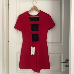 Röd kort klänning (jumpsuit/byxa under) öppen rygg med rosetter - Superfin röd kort jumpsuit med två stora rosetter backtill med öppen rygg. Klänningen har korta ärmar och en rak,elegant passform.