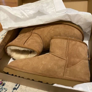 ugg minis - Mysiga bruna UGG boots i mjuk mocka med fårskinnsfoder. Klassisk rund tå och platt sula, perfekt för kalla dagar. Bootsens korta modell gör dem enkla att styla till både jeans och mjukisbyxor.