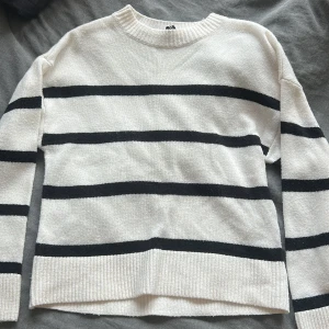 Vit och svart randig stickad tröja - Vit stickad tröja med svarta horisontella ränder och rund hals. Tröjan har långa ärmar och ribbade muddar vid hals, ärmslut och nederkant. Perfekt för dig som gillar en klassisk och stilren look.