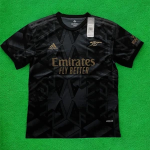 Arsenal svart bortatröja Adidas M - Snygg svart Arsenal bortatröja från Adidas med guldiga detaljer och tryck. Tröjan har korta ärmar, rund hals och ett geometriskt mönster i tyget. Materialet är lätt och andas, perfekt för fotboll eller streetwear. Emirates Fly Better-logga på bröstet.