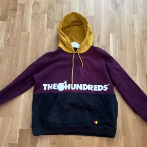 Colorblock hoodie från The Hundreds - Säljer en hoodie från The Hundreds i storlek L. Tröjan har färgblock i vinrött, svart och senapsgult, med stor vit logga fram och bak. Huva med snörning och känguruficka. Perfekt för dig som gillar streetwear och vill sticka ut.