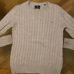 Grå kabelstickad tröja från GANT - Säljer en grå kabelstickad tröja från GANT i premium bomull. Tröjan är i gott använt skick och har rund halsringning, ribbade muddar och diskret GANT-logo broderad på bröstet. Det finns en fläck under höger armhåla. Man ser inte fläcken ifall man har på sig den. Perfekt för lager-på-lager och chill höst- eller vinterstil.