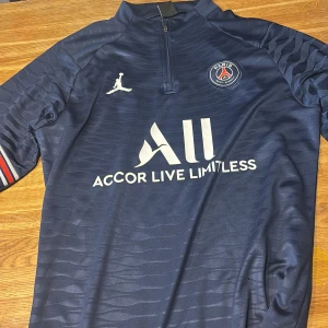 PSG långärmad träningströja Jordan - Mörkblå långärmad PSG träningströja från Jordan med halv dragkedja, vita loggor och texten 'Accor Live Limitless' på bröstet. Tunn, sportig polyester med diskret vågmönster och rödblå detalj på ärmen. Perfekt för fotboll eller träning.