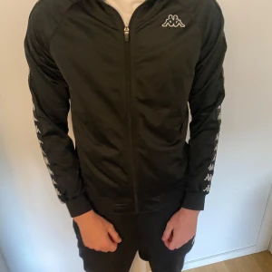 Svart Kappa vindjacka med loggor - Svart vindjacka från Kappa med klassiska vita loggor längs ärmarna och på bröstet. Använts flitigt men är fortfarande i bra skick! Säljer då den är för liten.