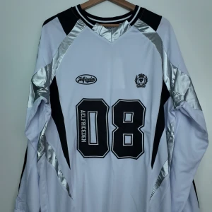 AELFRIC EDEN TRÖJA - Snygg vit långärmad fotbollströja från AELFRIC EDEN med svarta och silvriga detaljer. Stort tryck med siffran 08 och texten Aelfriceden på framsidan. Tröjan har sportig design med svarta paneler och silvriga inslag i axelpartiet. Tillverkad i lätt och ventilerande material. Tröjan är helt oanvänd och togs endast ut ur dens påse för att ta bilder på den. Pris kan diskuteras.