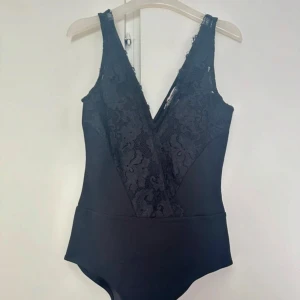 Svart spetsbody med v-ringning - Snygg svart body med breda axelband och djup v-ringning framtill. Framsidan har ett inslag av blommig spets som ger en lyxig känsla. Baksidan är enkel och slät. Perfekt att styla med jeans eller kjol för en trendig look
