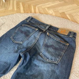 Blå jeans från Nudie Jeans, straight fit - Säljer ett par klassiska blå jeans från Nudie Jeans med raka ben och normal passform. Jeansen har snygga kontrastsömmar, fem fickor och Nudie-logga på bakfickan. Tillverkade i slitstarkt bomullsjeansmaterial.