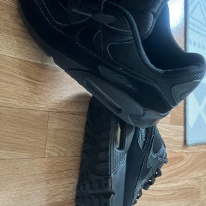 Svarta Nike Air Max sneakers - Säljer ett par helsvarta Nike Air Max 90 med klassisk chunky sula och synlig Air-enhet. Skorna har ovandel i mesh och skinn, med snygga detaljer och snörning framtill. Perfekta för dig som gillar en clean och sportig look. Typ helt nya.
