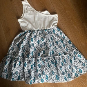 Vit och mönstrad oneshoulderklänning - Snygg kortklänning med vit, slät överdel i oneshoulder-modell och kjol med blått och svart grafiskt mönster på vit botten. Kjolen är volangig och ger en luftig känsla. Perfekt för dig som gillar unika detaljer och somrig stil.