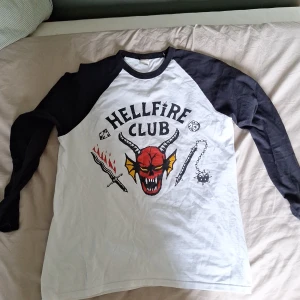 Hellfire Club långärmad tröja M - Säljer en långärmad Hellfire Club-tröja från Fruit of the Loom. Tröjan är vit med svarta ärmar och har ett stort tryck med demon, eldsvärd och tärningar på bröstet. Perfekt för dig som gillar Stranger Things eller vill sticka ut med en cool printad tröja.