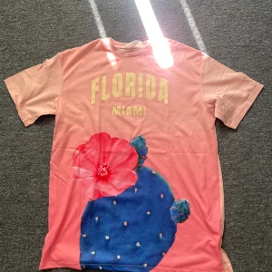 Rosa t-shirt med tryck från H&M - Snygg rosa t-shirt från H&M med stort tryck av blå kaktus och röd blomma framtill. Texten 'FLORIDA MIAMI' i gult på bröstet. Skön bomullskvalitet och klassisk passform med korta ärmar. Perfekt för dig som gillar färg och coola motiv!